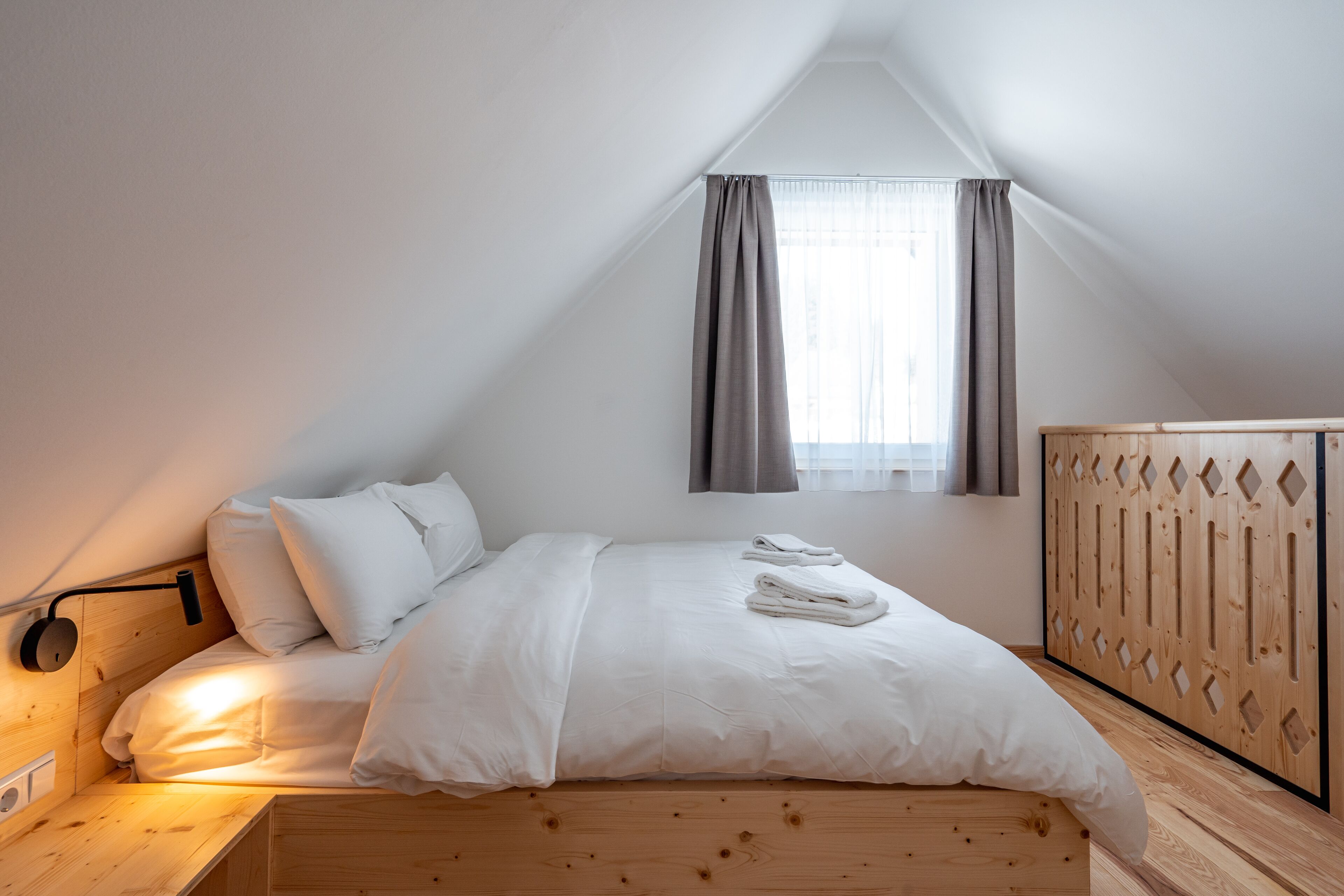 A bedroom with a wooden headboard, a bed with white linens, a bedside table with a lamp, and a window with curtains.