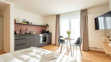 Ein modernes Hotelzimmer mit Bett, einer kleinen Küchenzeile, einem Esstisch mit Stühlen und einem an der Wand montierten Fernseher.