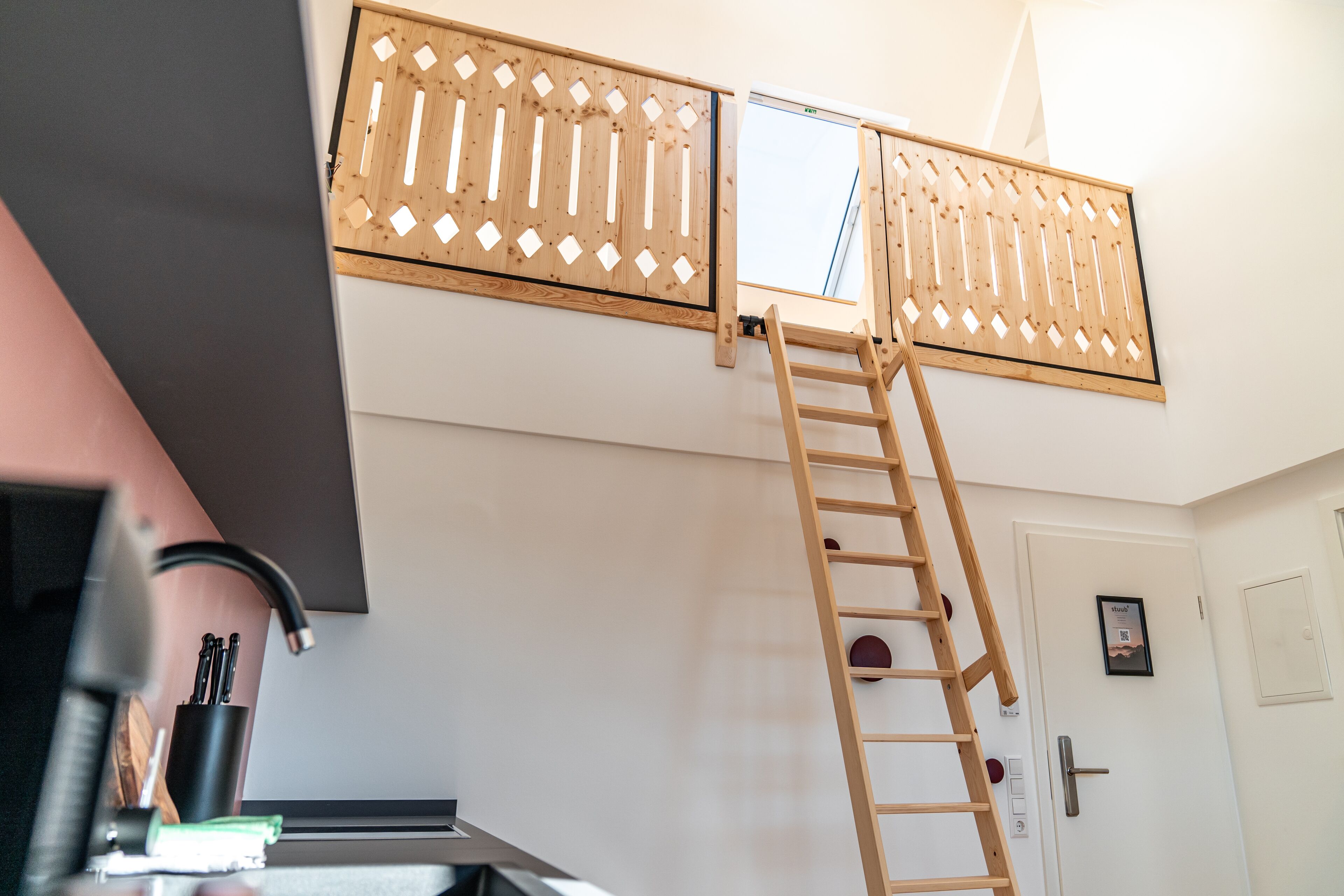 Een kamer met een houten ladder die leidt naar een vide met een raam, een deur met een fotolijstje, en een zwarte container met pennen op een bureau.