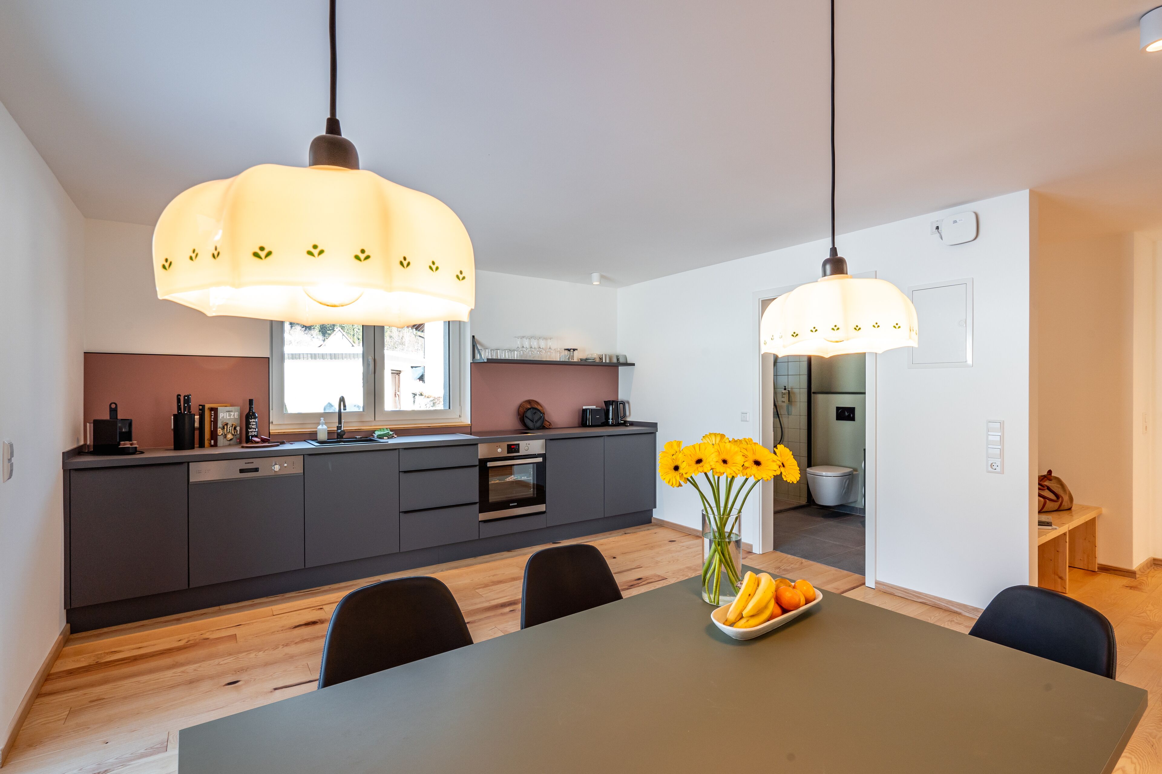 Eine moderne Küche mit dunklen Schränken, einer Holzarbeitsplatte und einem Essbereich mit Tisch und Stühlen. Zwei Pendelleuchten hängen über dem Tisch, und auf dem Tisch steht eine Vase mit Blumen.