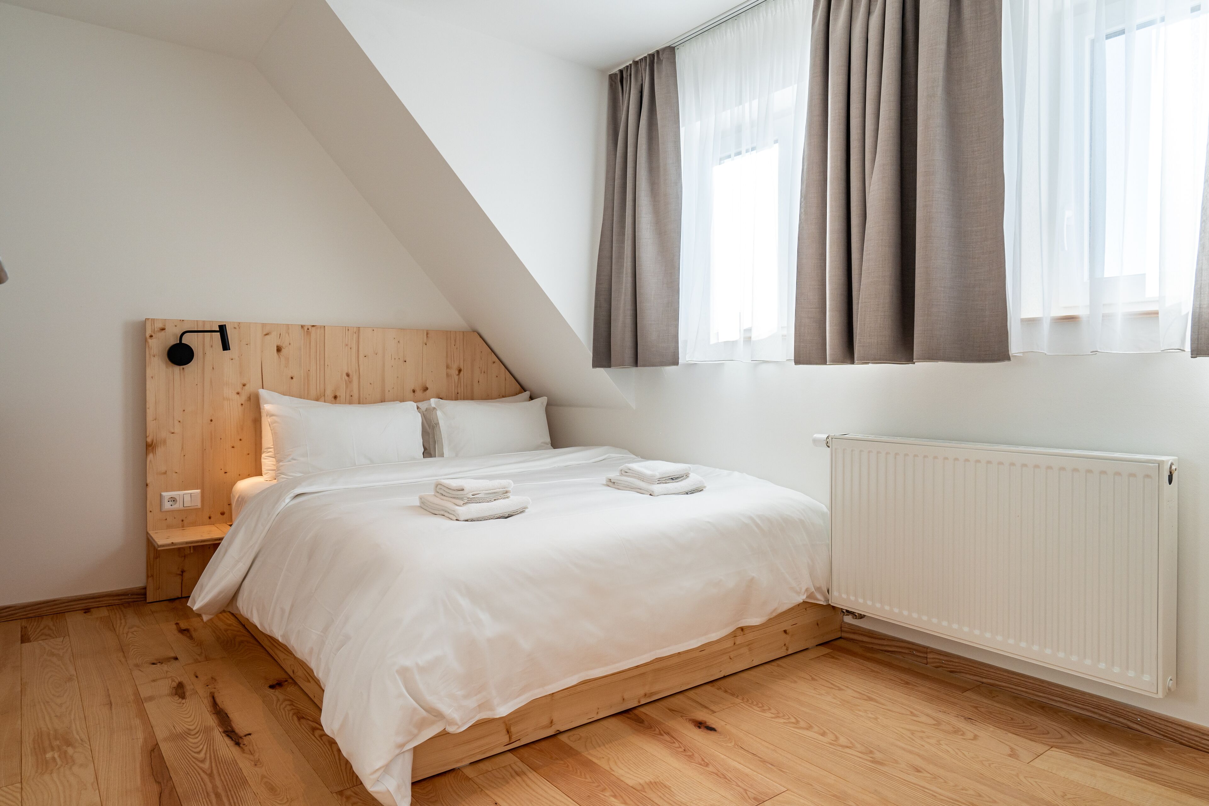 A bedroom with a sloped ceiling, a wooden headboard, a white bed with two pillows, and a radiator.
