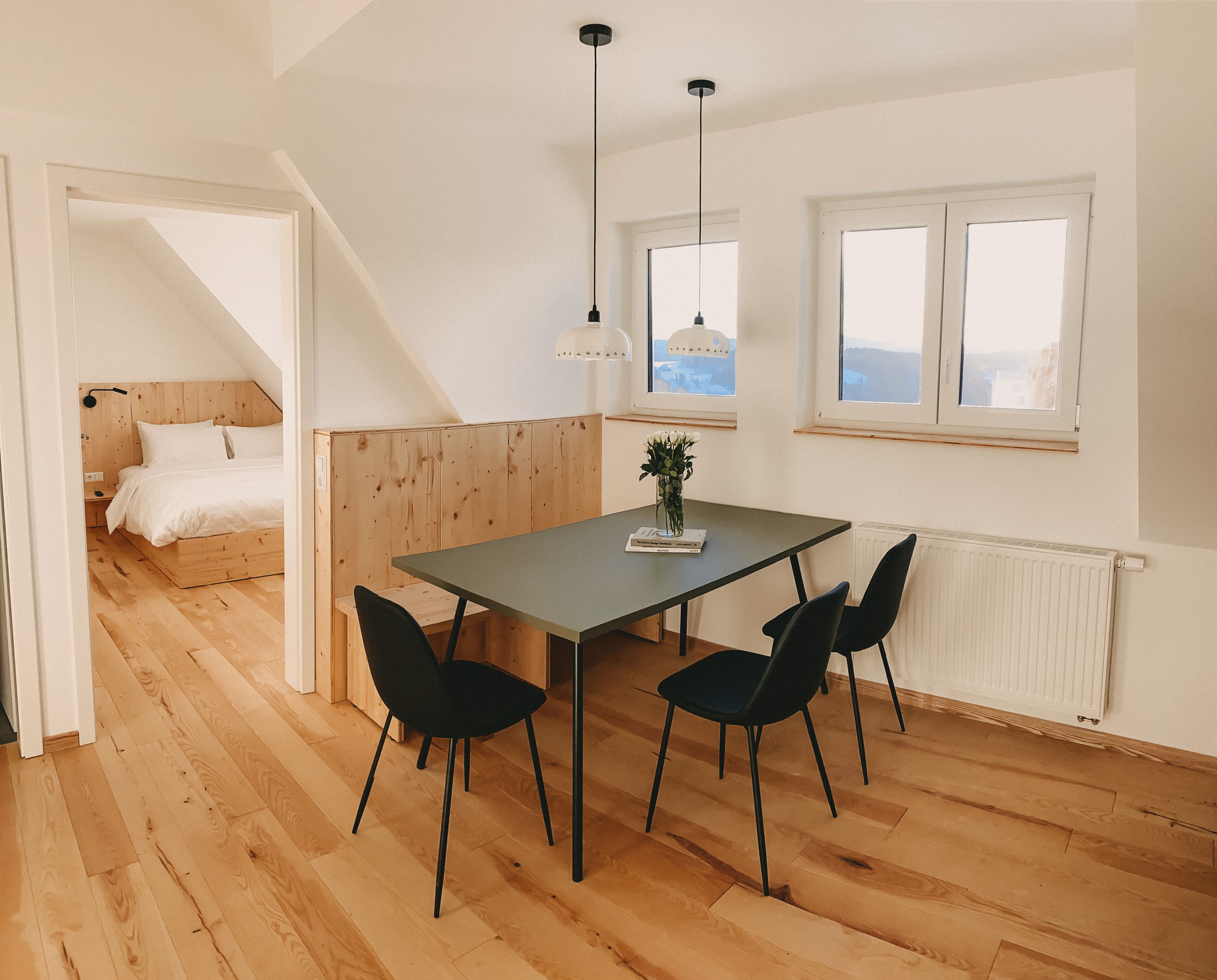Ein modernes Zimmer mit Holzboden, einem Esstisch mit Stühlen, einem Bett mit Holzvorzelt und einem Fenster mit Blick.