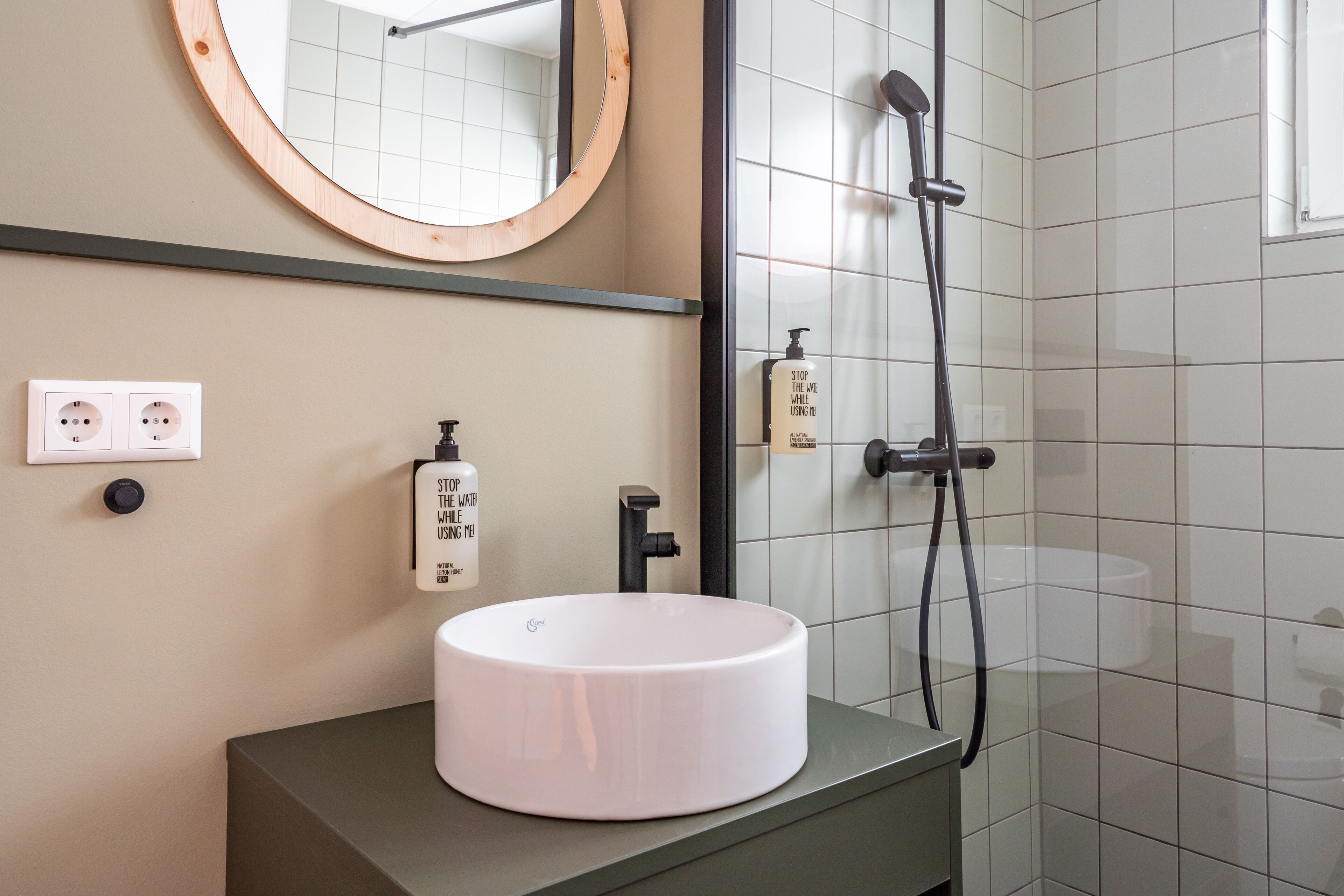 Une salle de bain avec un lavabo rond blanc, un miroir avec un cadre en bois, un robinet noir et une douche avec un tuyau noir.