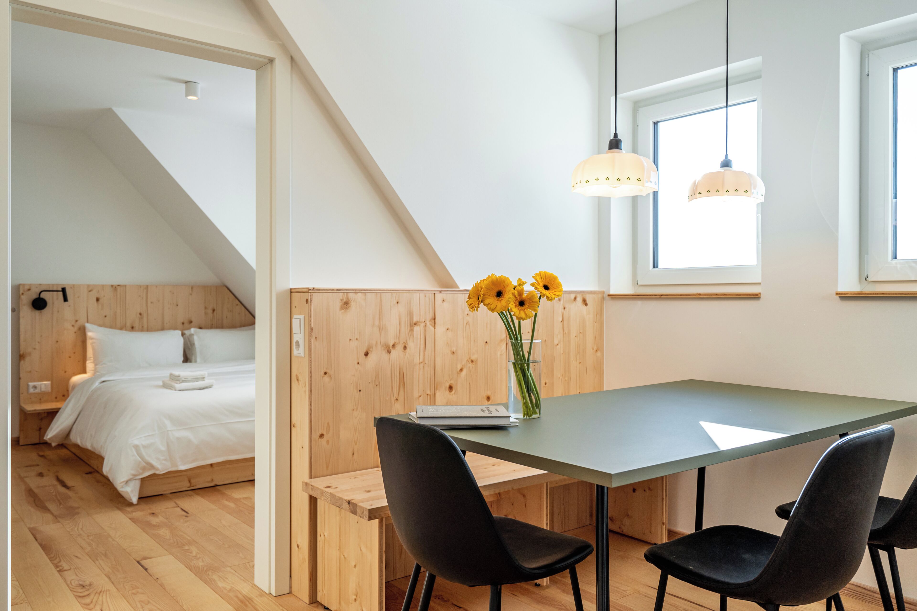Quarto moderno com cama, mesa com cadeiras, um vaso com flores e janela com luz natural.