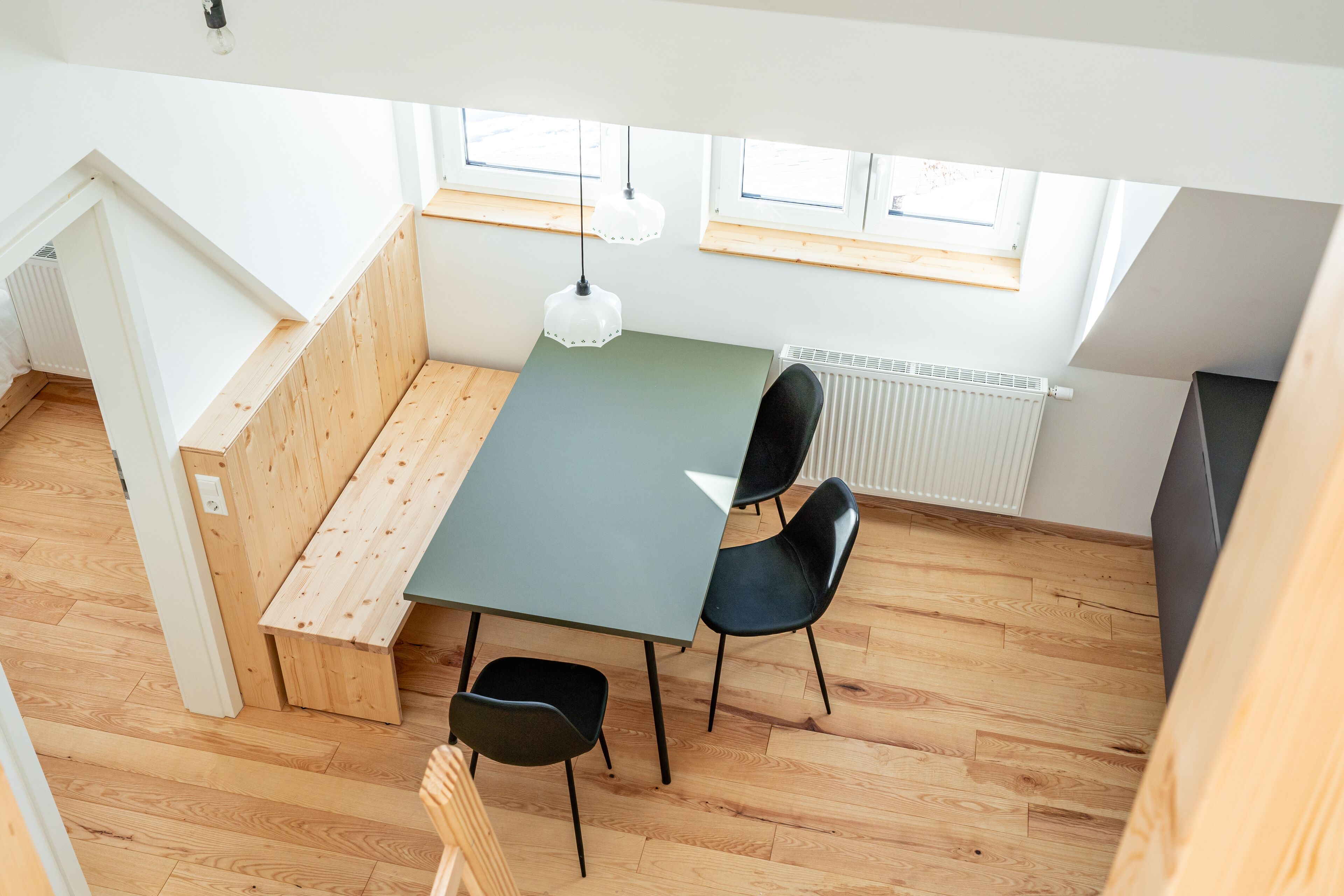 A modern, well-lit room with a wooden floor, a green table, and black chairs. There are windows with blinds and a radiator on the wall.