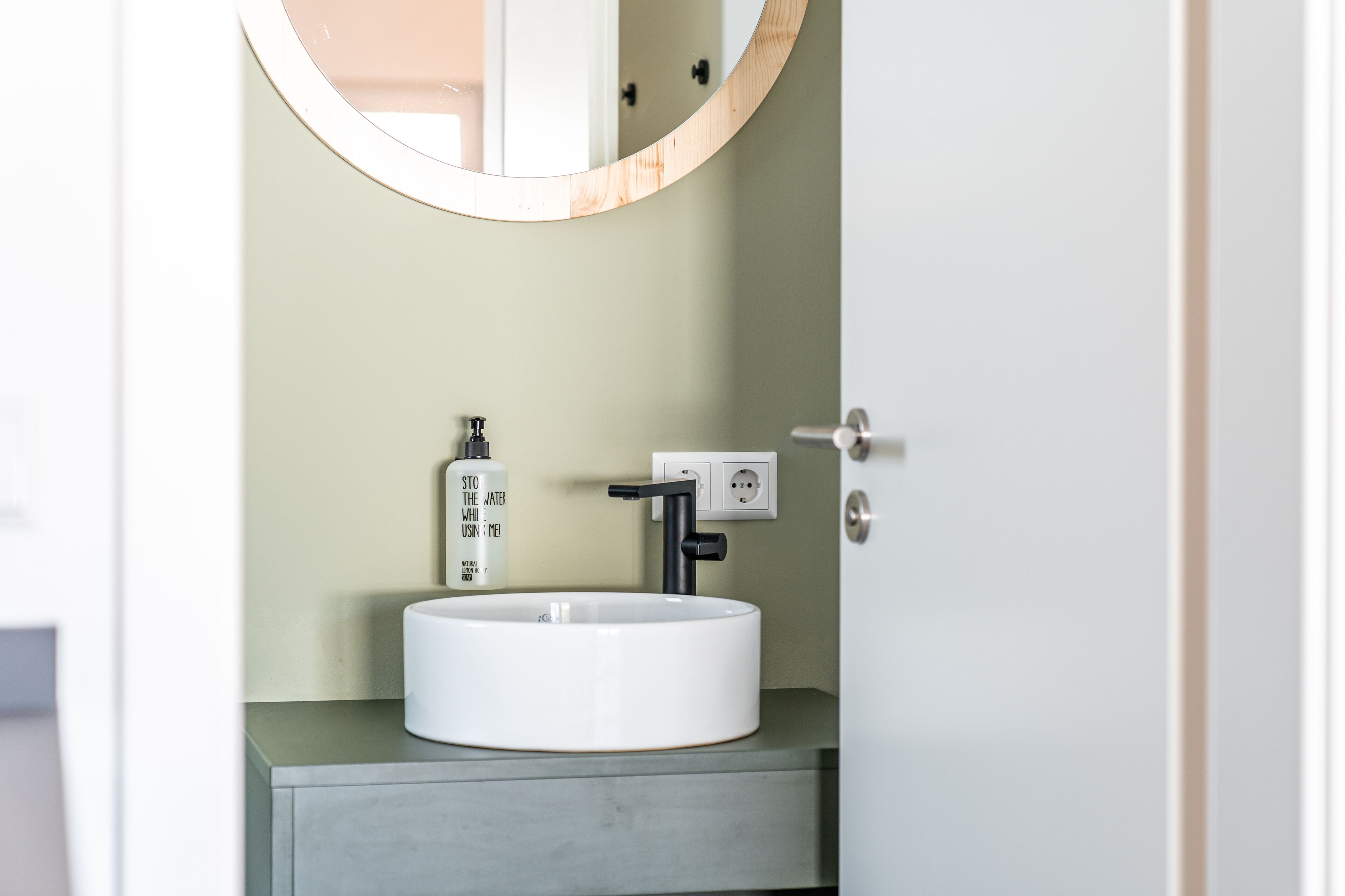 A bathroom with a round white sink, a black faucet, a mirror, and a bottle of hand soap.