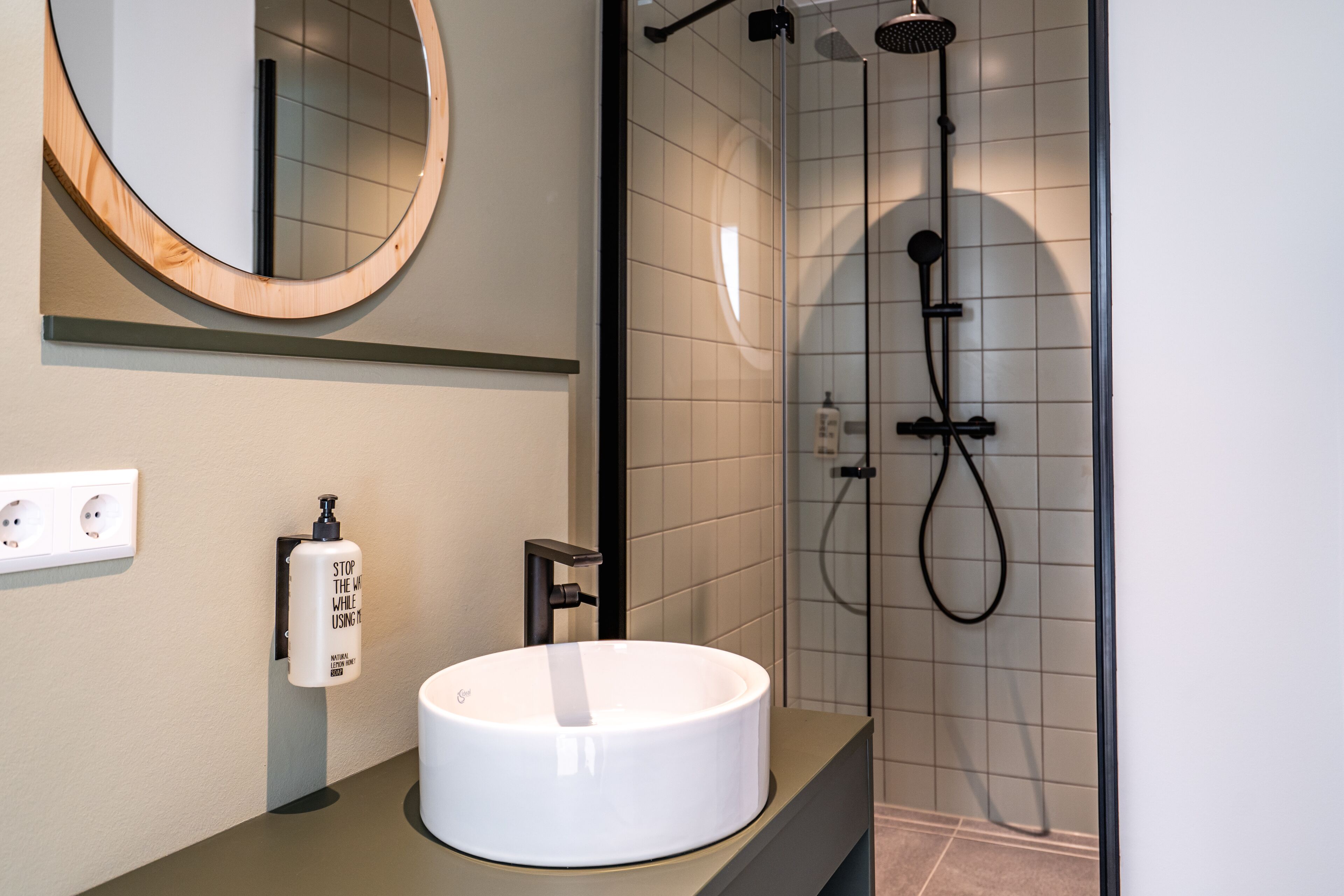 A bathroom with a round mirror, a white sink, a handheld soap dispenser, and a shower with a handheld showerhead.