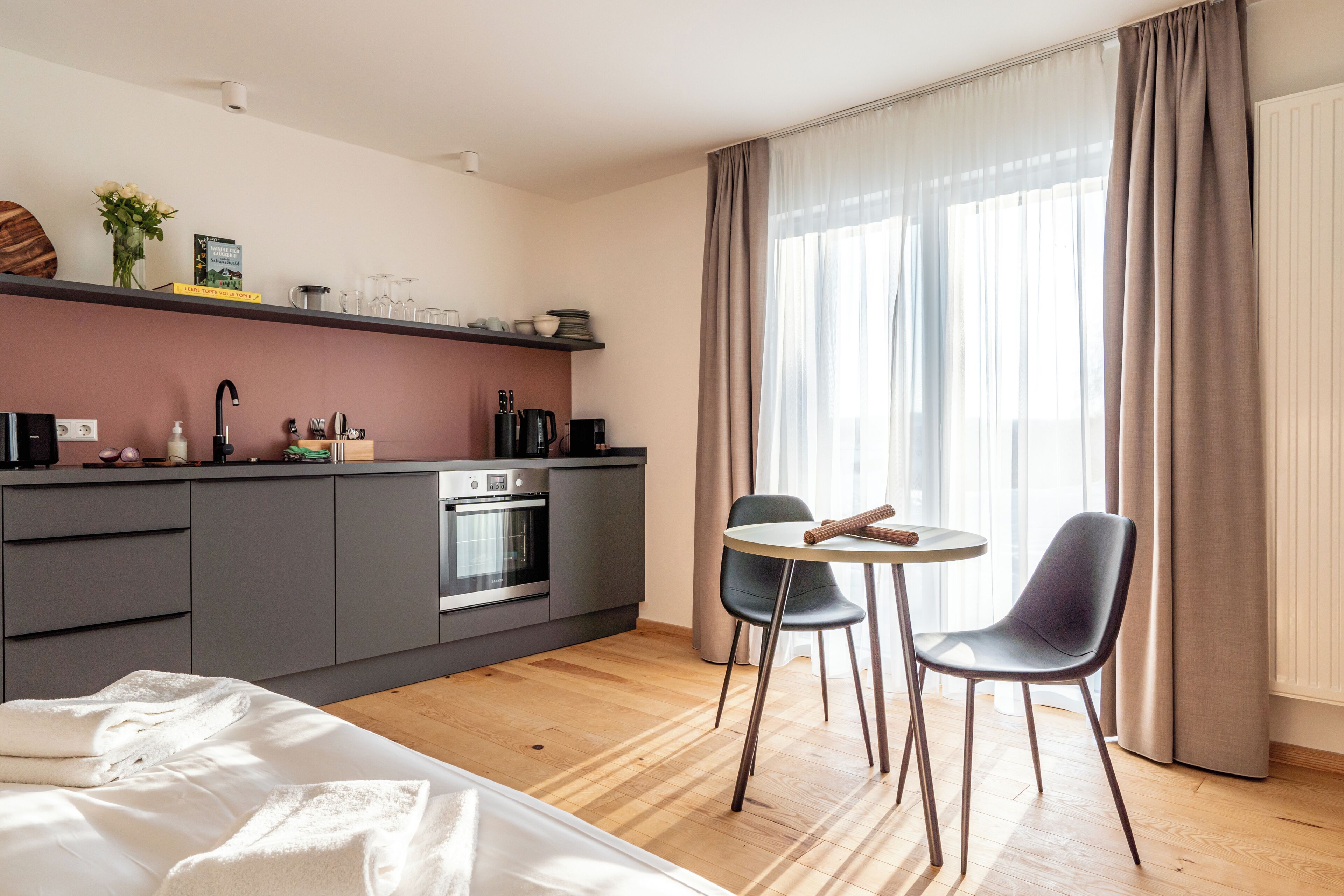 Una habitación de hotel moderna con kitchenette, cama, zona de comedor con dos sillas y un ventanal con cortinas.
