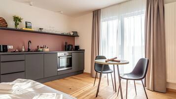 A modern hotel room with a kitchenette, a bed, a dining area with two chairs, and a large window with curtains.