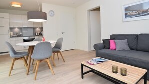 A modern living room with a grey sofa, a wooden coffee table, and a dining area with a white table and grey chairs. - fewo1846 - Smucke Utkiek (Glücksburg)