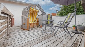 Une terrasse en bois avec une petite table et deux chaises, un banc en osier et un grand parasol.