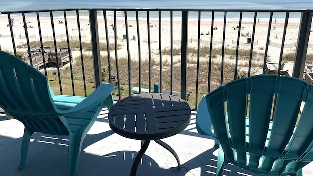 Un balcón con dos sillas turquesas y una mesita pequeña, con vistas a una playa donde hay gente y una terraza de madera.