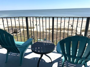 Ein Balkon mit zwei türkisfarbenen Stühlen und einem kleinen Tisch, mit Blick auf einen Strand mit Personen und einer Holzterrasse.