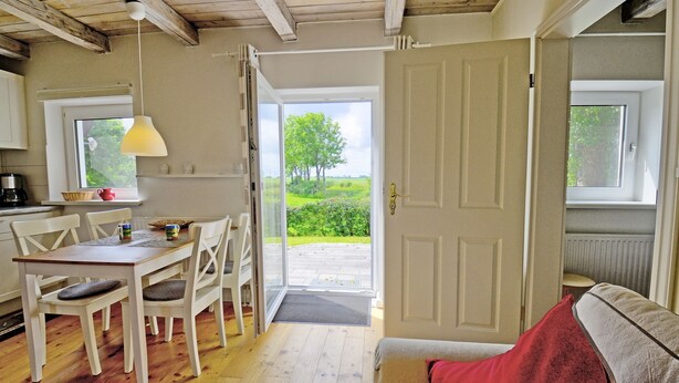 A cozy room with a wooden floor, a dining table with chairs, a sofa, and a window with a view of greenery. - fewo1846 - Red House - Holiday home with a covered terrace and 2 bedrooms (Husby)