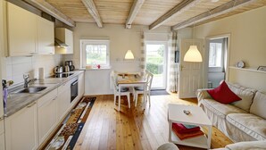 A cozy kitchen with wooden beams, a dining area with a table and chairs, and a living room with a sofa and a red pillow. - fewo1846 - Red House - Holiday home with a covered terrace and 2 bedrooms (Husby)