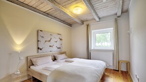 A bedroom with a wooden ceiling, a bed with white bedding, a wall-mounted artwork of animals, a window with curtains, and a radiator. - fewo1846 - Red House - Holiday home with a covered terrace and 2 bedrooms (Husby)