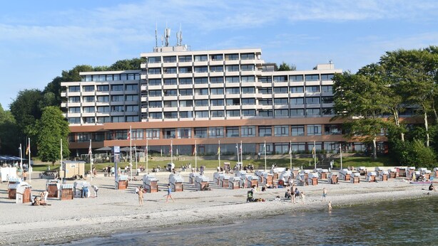 A large hotel with multiple floors, a beach area with umbrellas and sun loungers, and a body of water in the foreground. - fewo1846 - Kajüte Fördeblick (App. 112) / family-friendly studio apartment (Glücksburg)