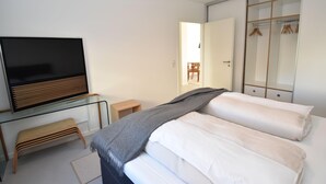 Ein modernes Hotelzimmer mit Flachbildfernseher, einer hölzernen Bank, einem Bett mit weißen Leinen und einem begehbaren Kleiderschrank mit Kleidung.