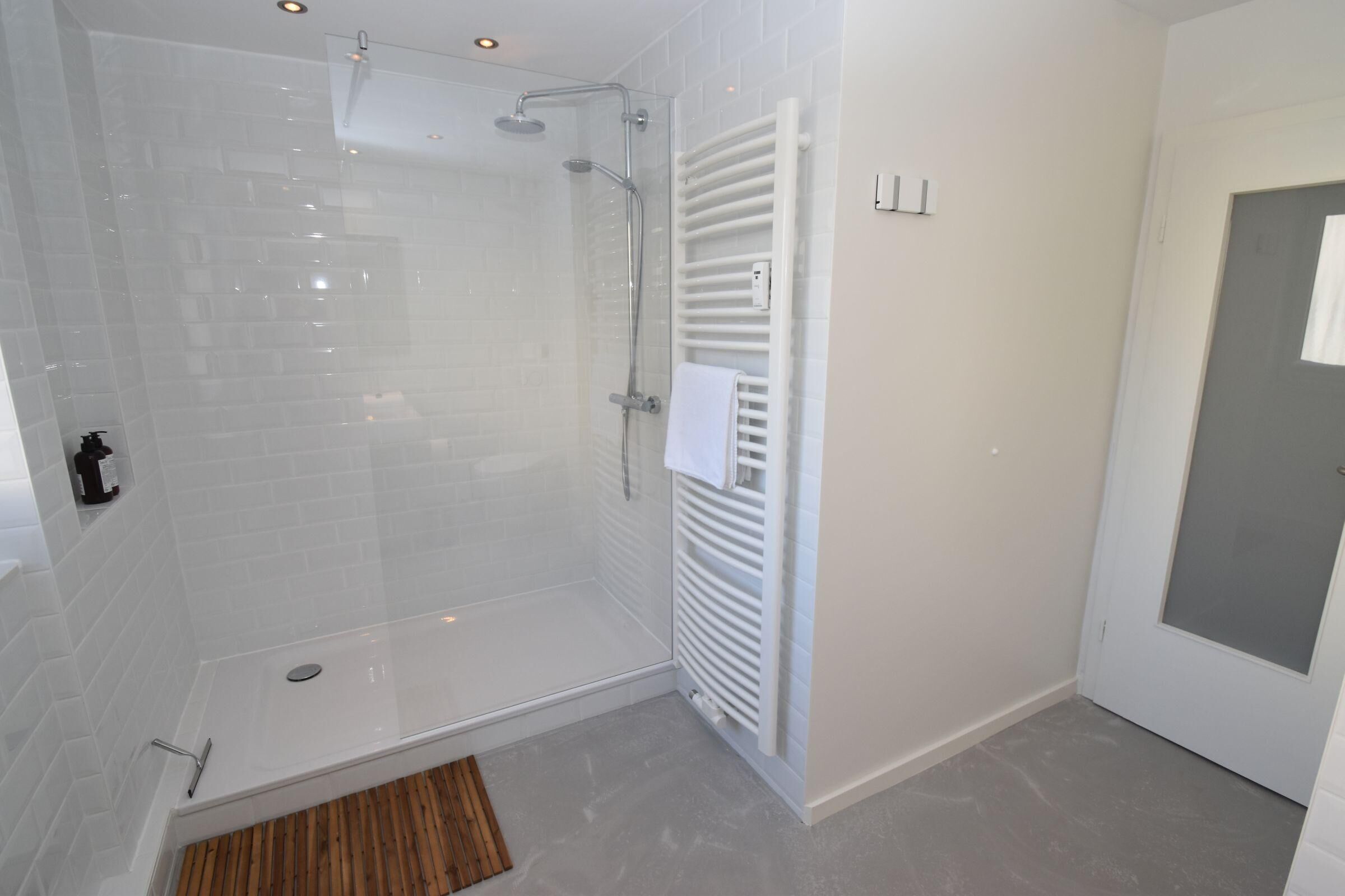 Un baño moderno con ducha de vidrio, toallero blanco y un tapete de madera en la entrada.