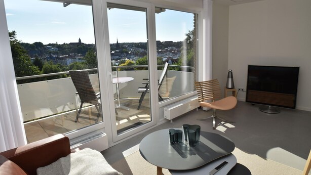 Ein modernes Wohnzimmer mit einem großen Schiebeglasfenster zum Balkon, einem Flachbildfernseher und einem Essbereich mit Tisch und Stühlen.