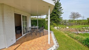 A patio with a table and chairs, a covered area with a white railing, and a view of a garden with a pond.