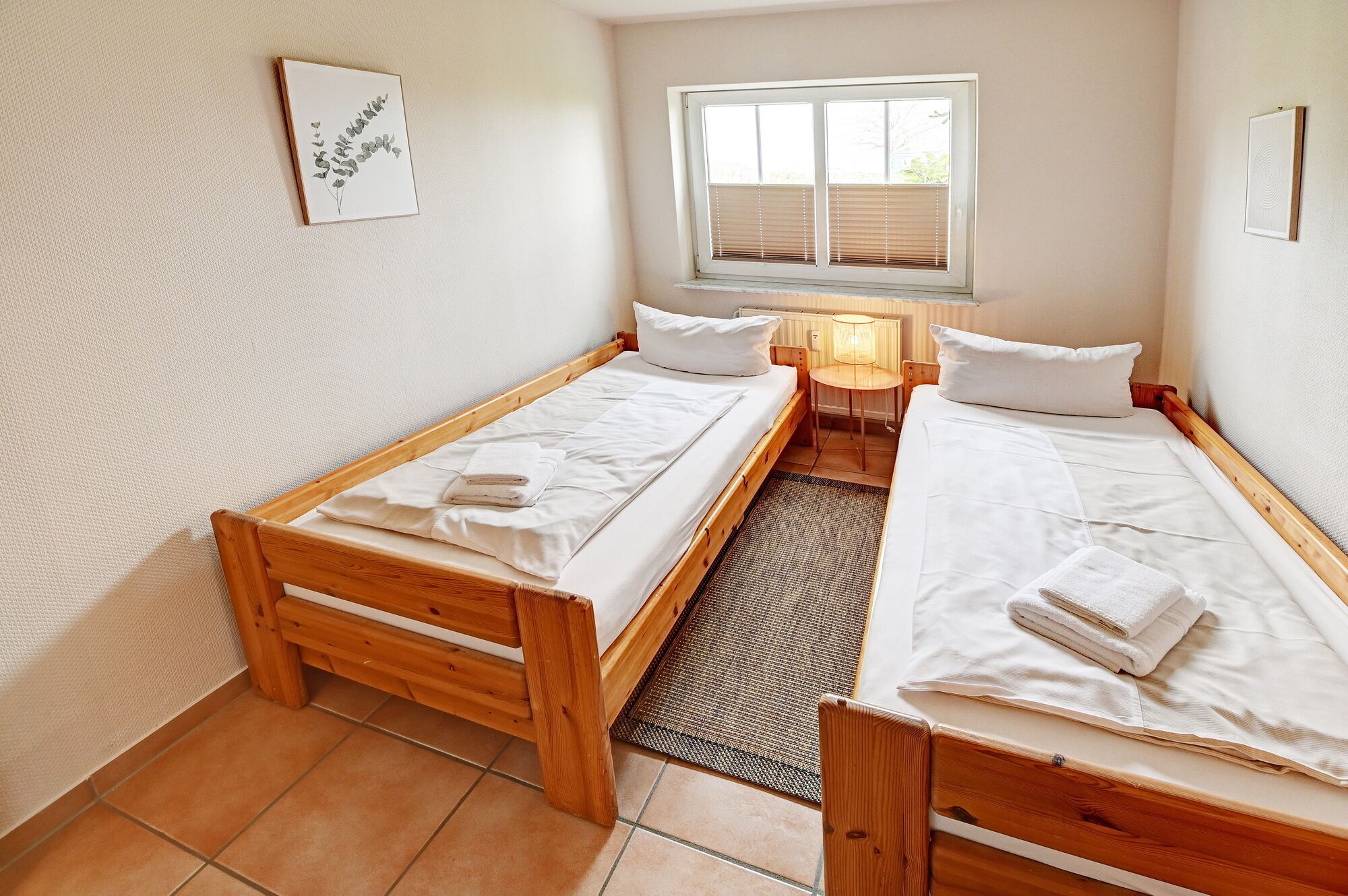 A small, clean room with two wooden beds, a window with blinds, a small table, and a rug.