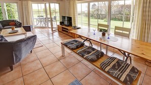 A spacious living area with a sofa, a dining table with benches, a TV, and large windows offering a view of the garden.