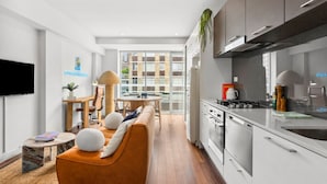 Een moderne keuken met witte kasten, een fornuis en een spoelbak. Er is een eethoek met een tafel en stoelen, en een bank met kussens. Een televisie is aan de muur bevestigd.