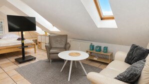 A modern loft bedroom with a sloped ceiling, a bed, a seating area with a sofa and armchair, a small round table, and a television. - fewo1846 - Little Mermaid (App. 666) / studio apartment in the attic (Harrislee)