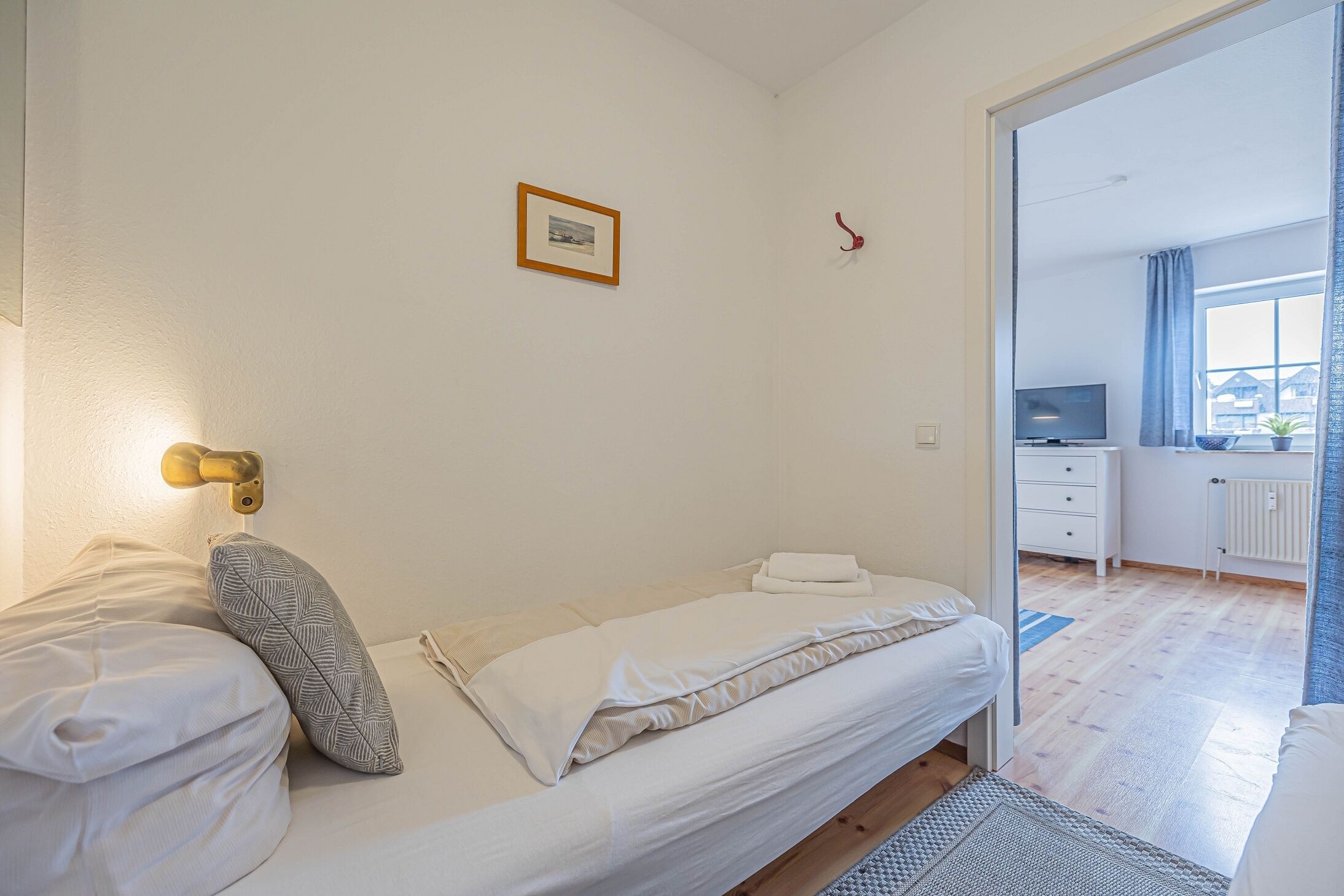 A bedroom with a bed, a bedside table, a small desk with a chair, a window with curtains, and a view of the outside.