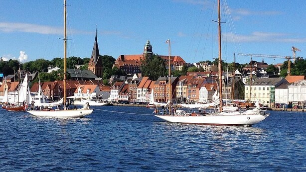 A harbor with several sailboats docked, a prominent church steeple, and a row of buildings along the waterfront.