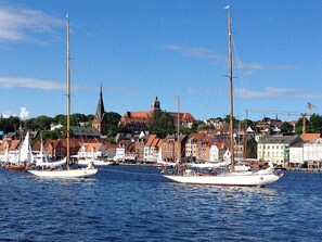 Ein Hafen mit mehreren Segelbooten an den Kais, ein markanter Kirchturm und eine Reihe von Gebäuden entlang der Uferlinie.