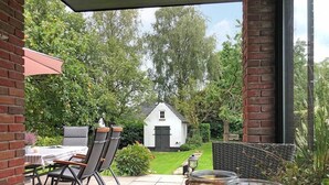 A patio with a table and chairs, a white building in the background, and a garden with trees and a lawn.