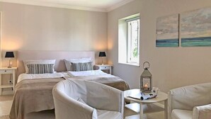 A bedroom with two beds, a nightstand with lamps, a small table with a lantern, and a view of the sea through a window.