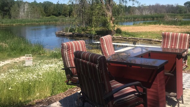 An outdoor seating area with striped chairs and a table, overlooking a lake and surrounded by trees.