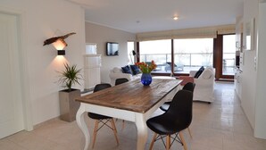 A modern living room with a wooden dining table, black chairs, a white sofa, and a TV mounted on the wall. - fewo1846 - Sundowner / Luxurious App. with 2 bedrooms, terrace, sea view (Flensburg)