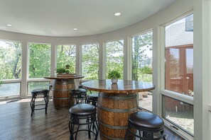 A sunlit room with a round wooden bar, stools, and large windows offering a view of greenery.