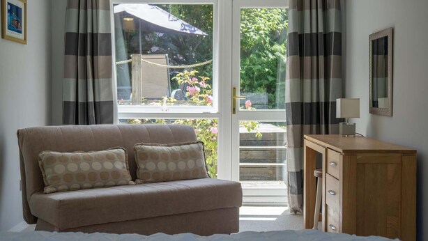 Ein Zimmer mit einem beige farbenen Sofa, einem Holzschrank und einer Glastür, die zu einem Außenbereich mit Sonnenschirm und Blumen führt.