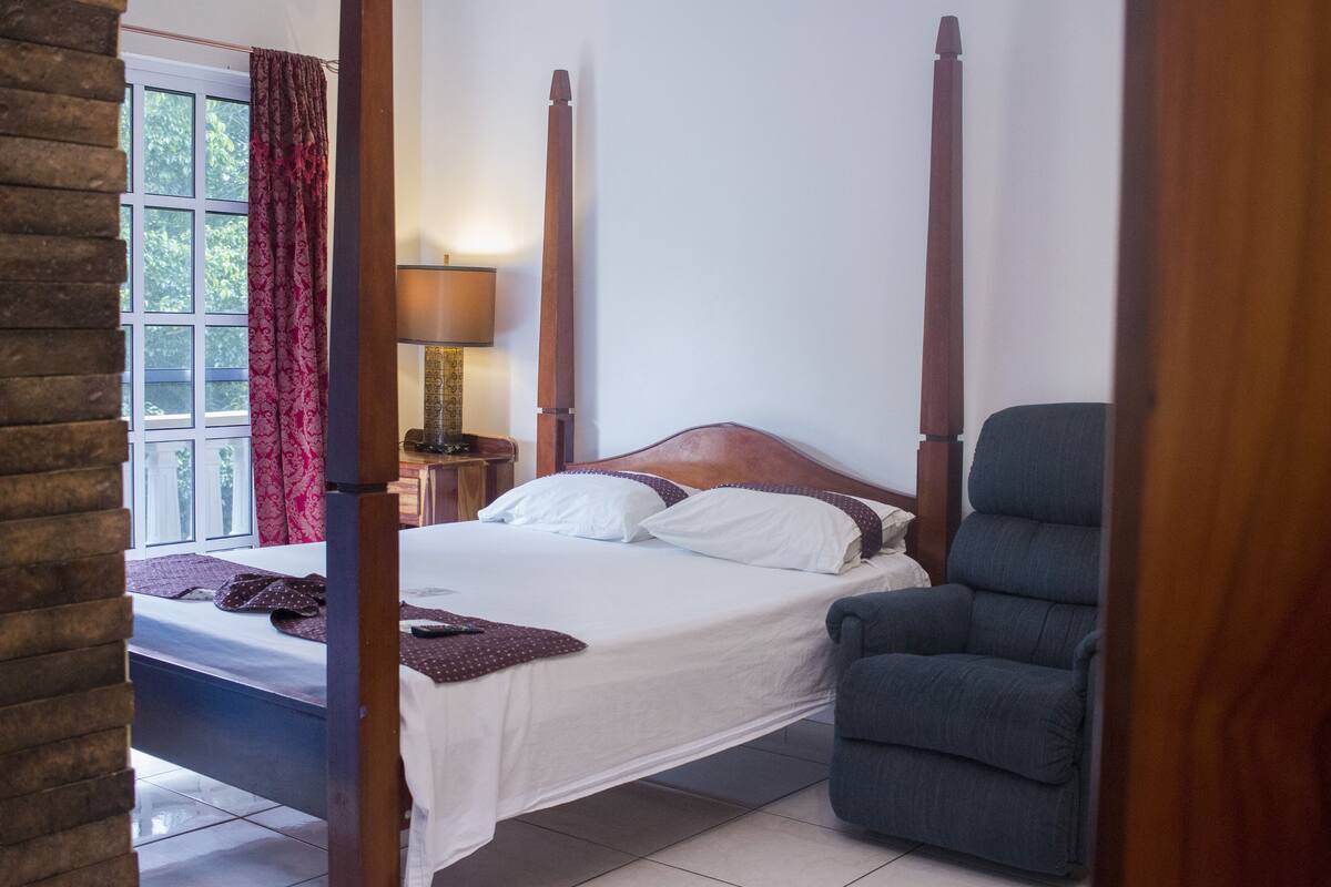 A four-poster bed with white linens, a blue armchair, and a bedside table with a lamp.