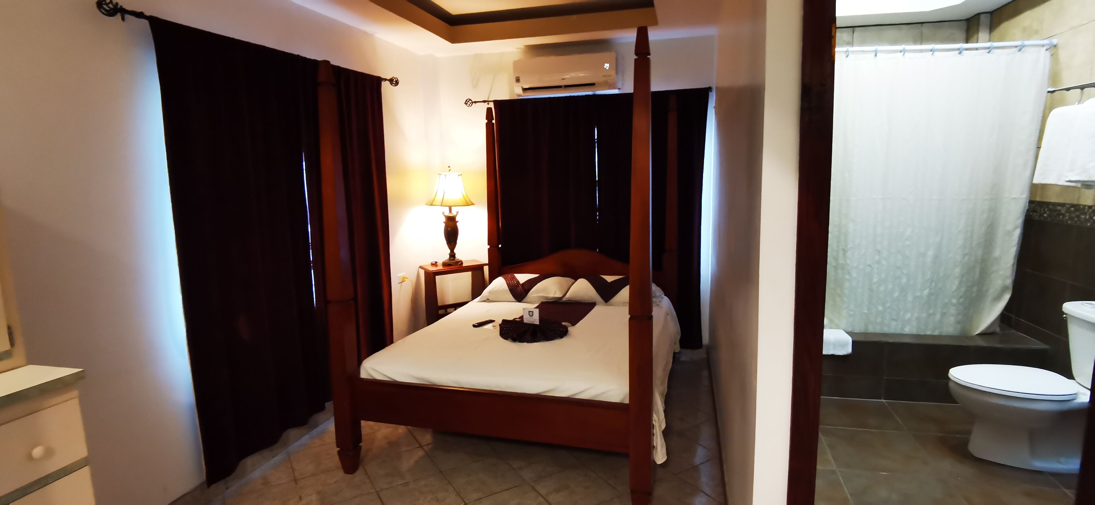 Une chambre d’hôtel avec un lit à baldaquin, une table de chevet avec une lampe, et une salle de bain avec douche et toilettes.