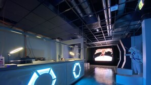 A futuristic lobby with a central reception desk featuring illuminated geometric patterns, a large digital screen displaying a space-themed video, and a statue of a space suit figure. - Space Night - Hostel (Berlin)
