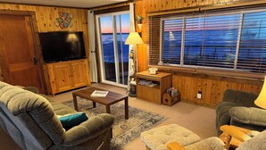 A cozy living room with wooden paneling, a television, a coffee table, and a view of the outdoors through large windows.