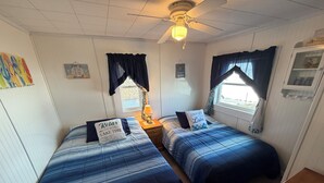 A room with two beds, a ceiling fan, a window with curtains, and a small table with a lamp.