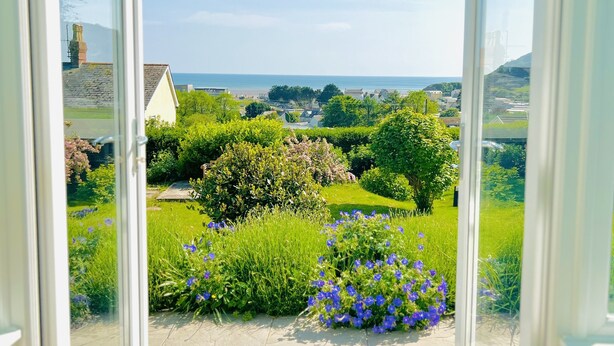 A view from a room with a large window overlooking a garden with various plants and a distant beach. - Seafacing Welsh cottage near the beach with stunning views accommodating 8.  (Pendine)