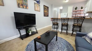 A living room with a flat-screen TV, a coffee table, two chairs, a sofa, and a kitchen area with a fridge and shelves.