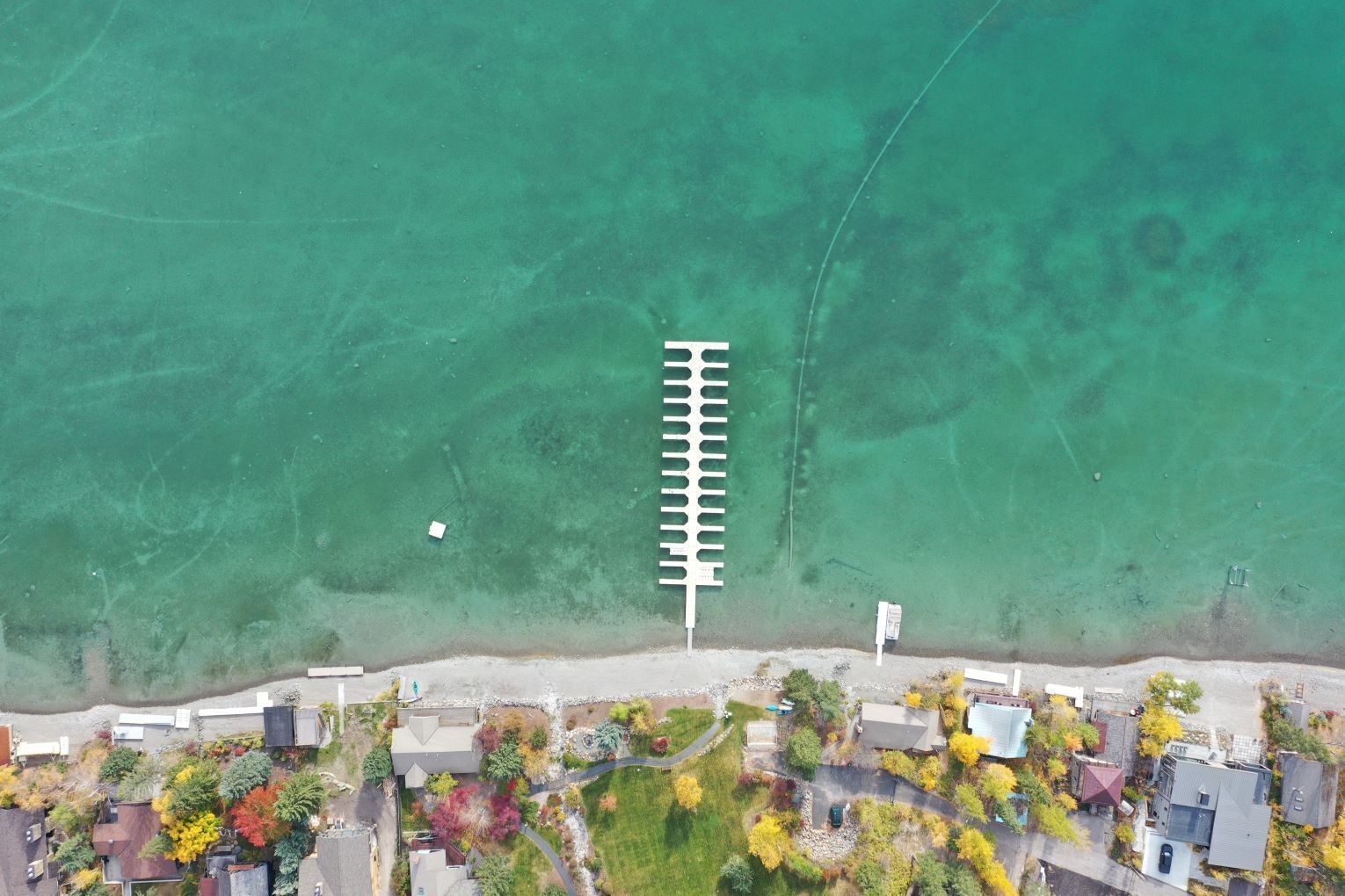Vue aérienne d’une propriété située sur la côte, avec une plage dégagée, une longue jetée blanche et différents bâtiments résidentiels dotés de jardins.
