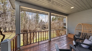 A covered porch with a wooden deck, a wicker chair, and a view of a wooded area. - Lakefront Property- Newly Updated Four Bedroom House, Sleeps 10 (Whitefish)