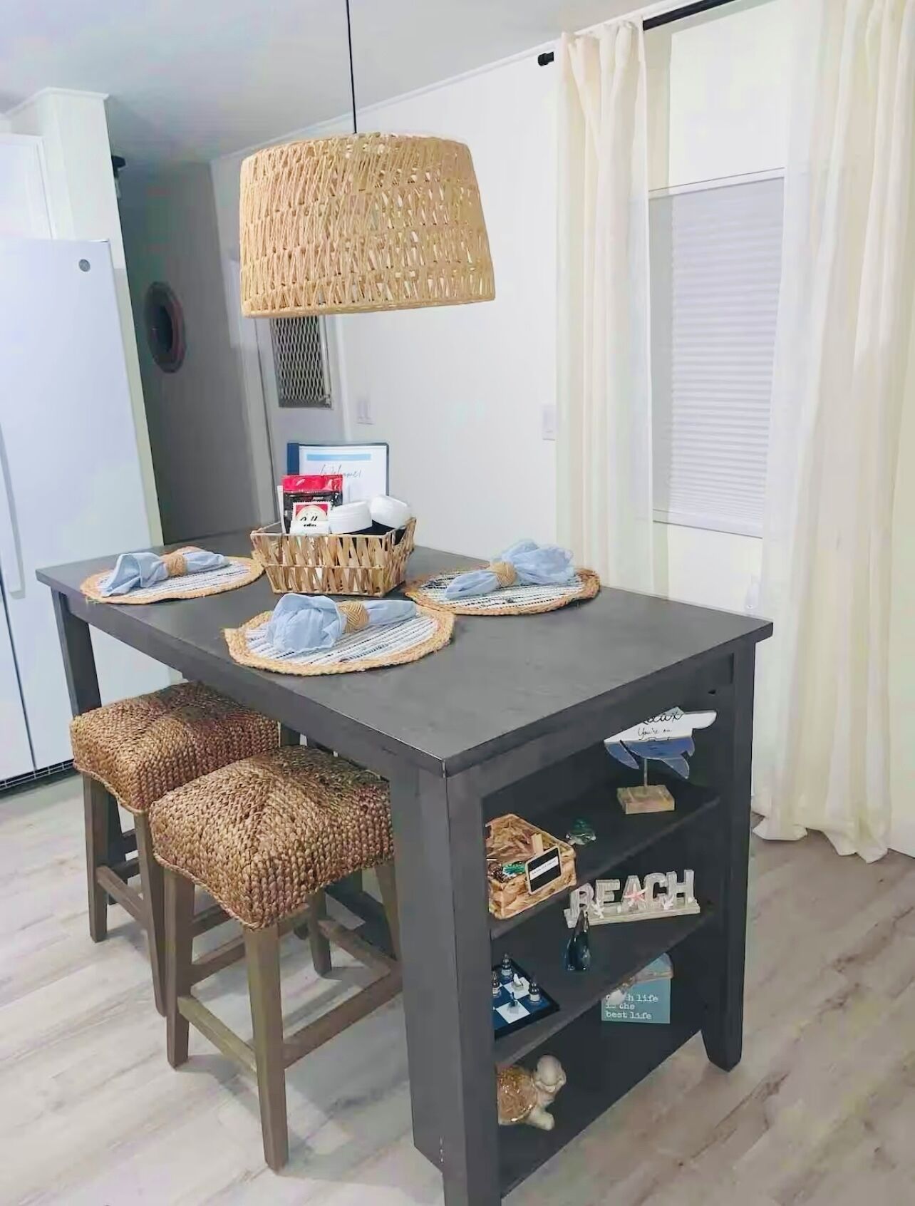 A dining table with woven placemats, two woven stools, a woven pendant light, and a shelf with decorative items.