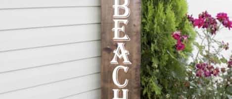 A wooden sign with the text "WELCOME TO OUR BEACH HOUSE" against a white house wall.