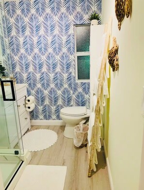 Et badeværelse med blå og hvid tapet med bladmotiv, et toilet, en glas brusekabine og en hvid kommode med spejl.